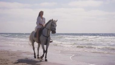 Güneşli bir günde plajda at süren beyaz elbiseli bir kızın geniş bir resmi.
