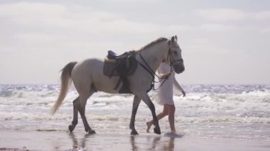 Beyaz bir elbise giymiş, güneşli bir günde atıyla kumsalda çıplak ayakla yürüyen bir kızın geniş bir resmi.