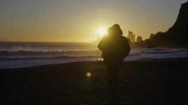 Reynisfjara sahilinin kara kumlarında yürüyen siluet batan güneşe ve uzaktaki Reynisdrangar deniz yığınına bakıyor.