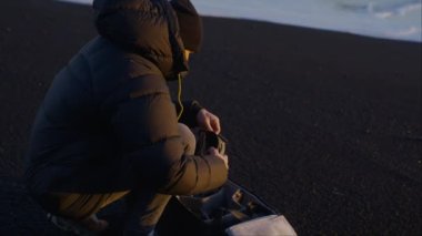 Kamera, İzlanda 'daki Reynisfjara plajındaki kara kumlara insansız hava aracını tamir ederken ceketini giyen bir adama yavaşça zumluyor.
