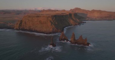 Reynisdrangar deniz yığınları ve Reynisfjall dağlarının İzlanda kıyılarının karanlık kumlarıyla tezat oluşturduğu Reynisfjara 'nın çarpıcı manzarasının üzerinde bir dron uçuyor.