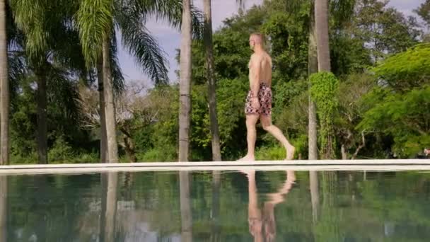 Muž Pestrobarevných Trenýrkách Kráčí Podél Bazénu Lemovaného Palmami — Stock video