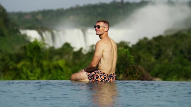 Man Trunks Sunglasses Looks Admires Iguazu Falls Distance While Sitting — Stock Video