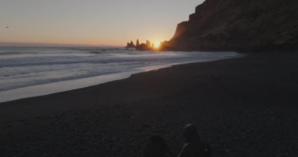 一架低空飞行的无人驾驶飞机飞越了站在雷尼斯法哈拉海滩黑色沙滩上的两名男子 飞机在日落时接近雷尼斯兰加岩层 — 图库视频影像