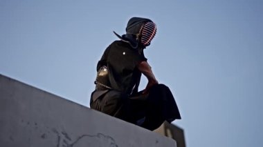 Kendo kıyafetli bir adam sakin bir varlıkla oturur, aşağıdaki kentsel manzaraya bakar.