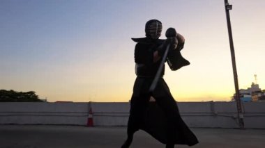 Kendo kıyafeti giyen bir adam gün batımında bir çatıda kılıcını sallarken etrafında döner ve yuvarlanır.