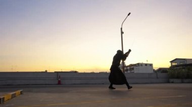 Kendo kıyafeti giyen bir adam gün batımında çatıda takılır ve kılıç tekniklerini uygular.