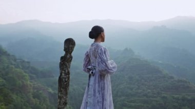 Lavanta dantelli bir elbise giymiş, taş bir sütunun yanında dikilmiş, mavi saat boyunca pirinç teraslarını ve dağları seyreden bir kadının geniş bir görüntüsü.