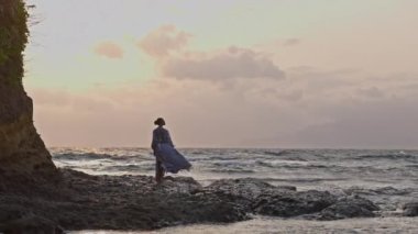 Elbiseli bir kadın kıyıya çarpan deniz dalgalarını seyrediyor.