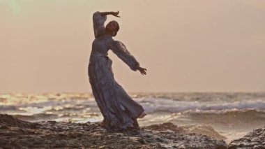 Gün batımında deniz dalgalarına karşı dokunaklı el hareketleriyle dans eden bir kadın.