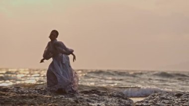 Dantel elbiseli bir kadın gün batımında dalgalara karşı kayalık sahilde dans ediyor.