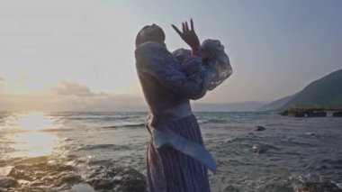 Gün batımında dalgalı bir deniz kıyısında danseden dantel bir elbise giymiş bir kadının orta boy fotoğrafı.