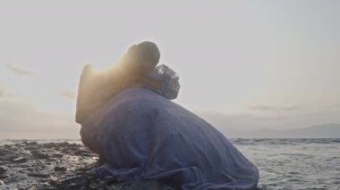 Bir kadın huzur içinde oturur ve kendi düşüncelerinde kaybolmuş kayalık bir sahilde teselli bulur.