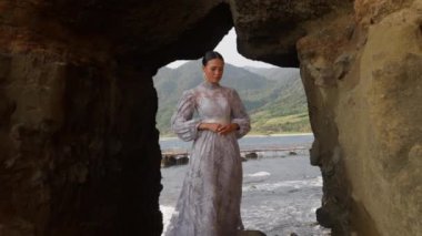 Lavanta dantelli bir elbise giymiş bir kadının, kayalık bir uçurumun üstünde, arkasında deniz olan bir kadının yakınlaştırılmış görüntüsü.