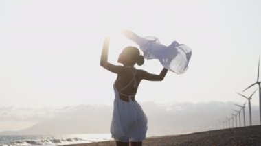 Gündüz vakti rüzgar türbinleriyle bir deniz kıyısında esen rüzgarı hissederken bir kadının arka görüntüsünü gösteren orta boy bir fotoğraf.