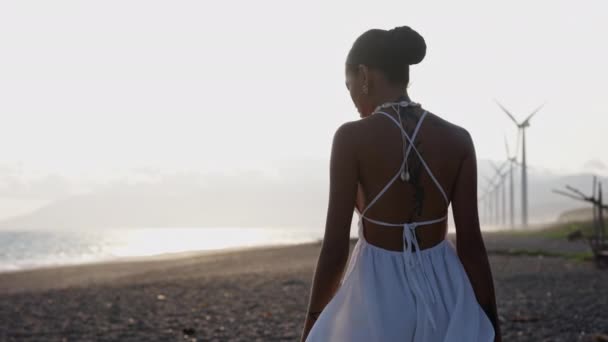 Beyaz Elbiseli Eşarbını Sağ Elinde Tutarken Rüzgar Türbinleriyle Sahil Kıyısında — Stok video