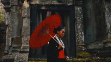 Elinde kırmızı bir şemsiyeyle siyah kimono giymiş bir kadın, gün içinde Kamboçya 'da terk edilmiş bir tapınağı keşfediyor.