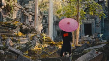 Elinde canlı kırmızı bir şemsiye tutan siyah kimonolu bir kadın, gündüz vakti Kamboçya 'da yosun kaplı bir tapınağı keşfediyor.