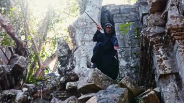 Siyah pelerinli maskeli bir adamın gündüz vakti terk edilmiş bir tapınakta büyük bir taş yığınının üzerinde durup kılıç duruşları yaparken geniş açılı görüntüsü.