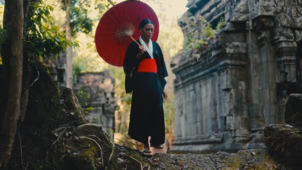 赤い傘を手に入れた黒い着物の女性は 昼間のカンボジアの放棄された寺院を歩きます — ストック動画