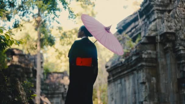 黒い着物の女性のバックビューショット 赤い傘を手に 昼間のカンボジアの放棄された寺院を探索 — ストック動画