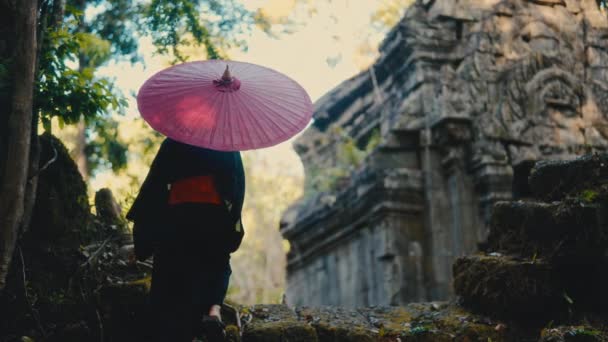 黒い着物に赤い傘を手に入れた女性のバックビューショットは 昼間にカンボジアで放棄された寺院を見るために 化石の石を登ります — ストック動画