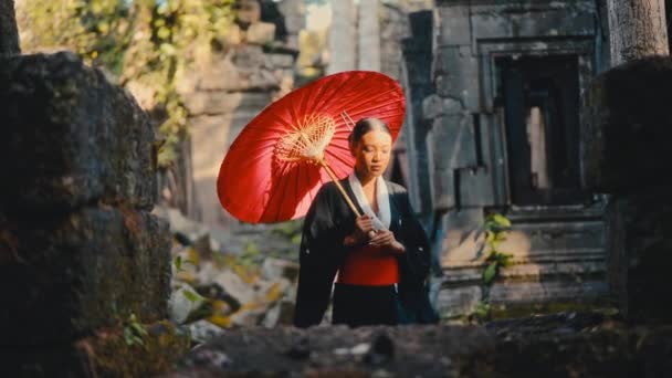 黒い着物を着た女性は 昼間にカンボジアの放棄された寺院を散策しながら赤い傘を運ぶ — ストック動画