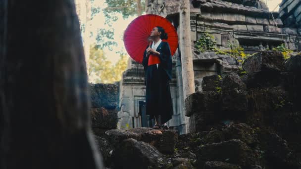 黒い着物を着た女性は 昼間にカンボジアで放棄された寺院を見ながら赤い傘を運ぶ — ストック動画