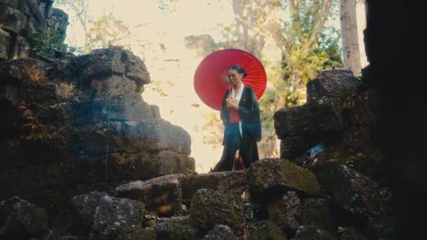 黒い着物を着た女性は 赤い紙の傘を持って 化石の石を登り カンボジアの放棄された寺院を見回します — ストック動画