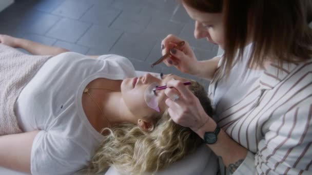 一位年轻女子躺在床上的头像 一位美容师用一把金属镊子在美容院里用睫毛膏展开 — 图库视频影像