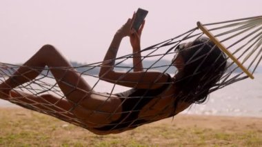 Mayo giymiş bir kadın hamakta güneşlenirken Koh Mak 'ta bir plajda telefonunu kontrol ediyor.