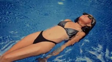 Şık bir kadın, güneş gözlüğü takmış, zahmetsizce havuzda yüzüyor.