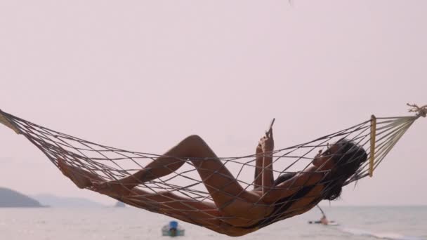 海辺の静けさの瞬間を象徴する 女性はコメのビーチでハンモックにリクライニング ソーシャルメディア接続とのリラクゼーションのバランスをとる — ストック動画