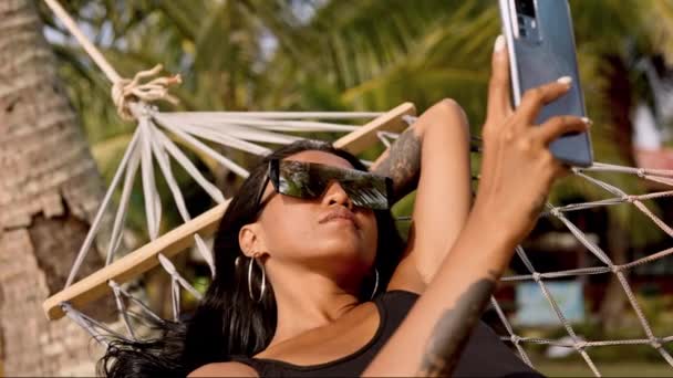一位时髦的女士戴着太阳镜 舒舒服服地躺在吊床上 沉溺于智能手机时代 享受着终极休闲 — 图库视频影像