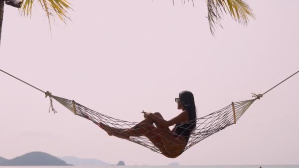 一名妇女躺在吊床上 在海滩边检查手机 沐浴在温暖的阳光下 — 图库视频影像