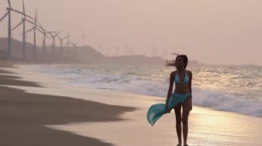 Mavi bikinili ve saronlu bir kadının, arka planda rüzgar türbinleri ve çarpan dalgalarla kumsalda yürüdüğü yavaş çekim.