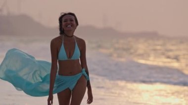 Mavi bikinili genç bir kadın ve sarong yavaş çekimde sahilde yürüyor ve kameraya yaklaşırken gülümsüyor.