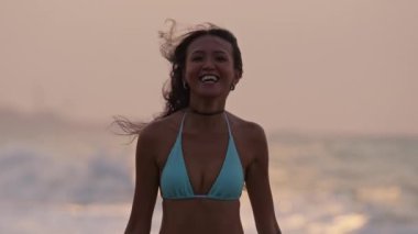 Plajda gün batımında kameraya gülümseyen mavi bikinili bir kadının orta boy yakın çekimi.