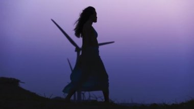 Sarong giymiş bir kadının yan görüntüsü, yürür ve kolları açık poz verir, alacakaranlıkta bir sıra rüzgar türbininin arasında.