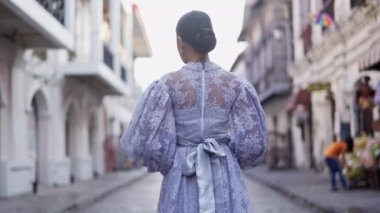 Şık çörek saç stili ve leylak rengi elbisesi olan bir kadın, Vigan 'ın canlı caddelerinde zarif bir şekilde geziniyor, kendini şehrin ebedi güzelliğine daldırıyor.