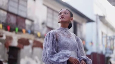 Tarihi bir caddede dikilen kahverengi saç stili ve leylak dantelli bir elbise giymiş bir kadın, kaldırım taşları ve sömürge mimarisi birleşerek zamansız bir güzellik ortamı yaratıyor.