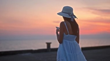 Akıllı telefonuna dalmış bir kadın iskelede dikiliyor, resimli bir günbatımının arkaplanına karşı.