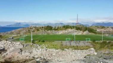 Soldan sağa hareket eden bir çekim, uzak dağ sıraları tarafından çerçevelenmiş bir adada resim gibi bir futbol sahasını gösteriyor.