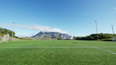 Bir futbol sahasının yapay sahası ve gündüz vakti uzak bir dağ manzarasıyla çekilen bir çekim.