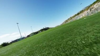 Bir İHA, açık mavi gökyüzünün altında bir futbol sahasının üzerinde uçuyor.