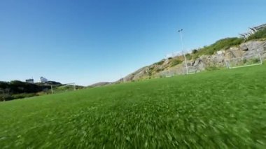 Geniş futbol sahasının üzerinde uçan bir İHA açık mavi gökyüzünün altında yapay çim