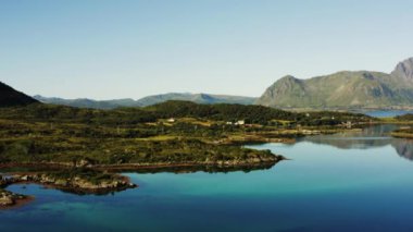 Canlı yeşil manzaralar ve berrak mavi gökyüzünün altındaki görkemli dağ zirveleri ile çevrili pitoresk bir göl üzerinde drone.