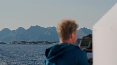 Mavi kapüşonlu ceketli bir film yapımcısının, feribota binerken, nefes kesici deniz ve dağ manzarasını çekerken arka plan görüntüsü.
