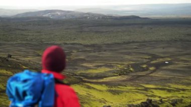Ceket ve sırt çantası giymiş, Raudhaskal kraterini çevreleyen karanlık manzarayı kaplayan yeşil yosun tabakalarına bakan bir adamın arkasından çekilen fotoğraf.