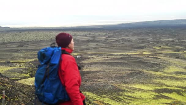 ジャケットの男とバックパックで 地平線上の明るい空を眺めながら ヒスラ火山の近くの黒と緑の風景を眺める — ストック動画